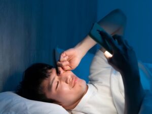 Man rubbing left eye and using phone while lying on bed